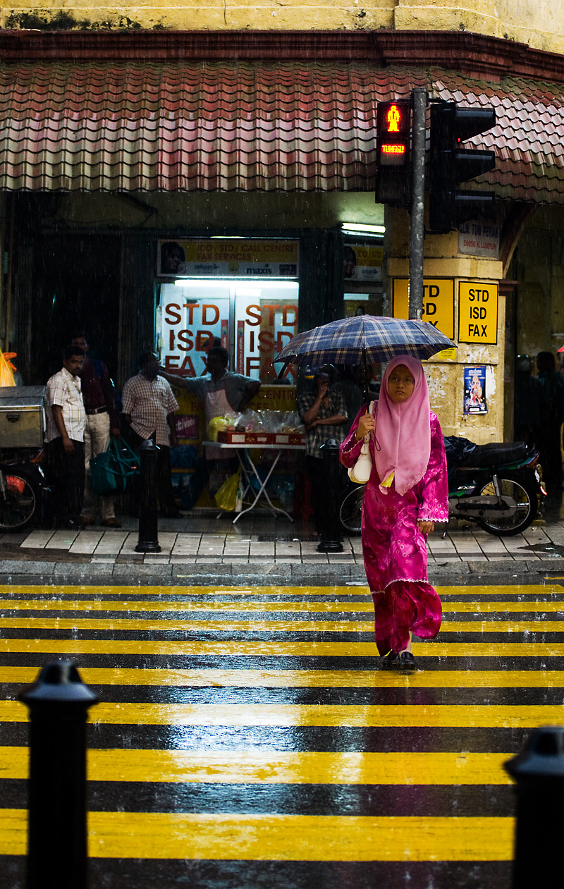 A Malaysian woman dressed in red clothes and pink head scarf crosses a striped street in the rain covered by a plaid umbrella. - Kuala Lumpur, Malaysia - Daily Travel Photos