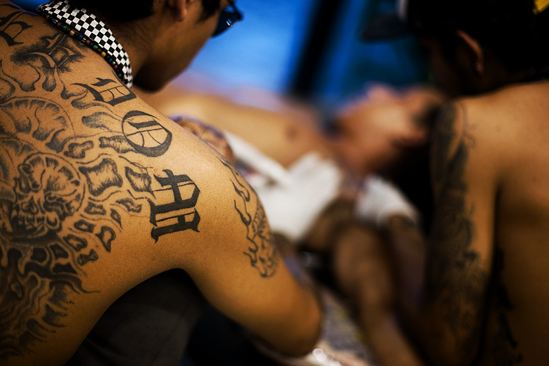 Two tattoo technicians draw a tattoo on a young traveler. - Ko Lipe, Thailand - Daily Travel Photos